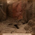 A narrow Arizona box canyon with steep red sandstone walls and a dark fissure opening in the canyon floor where horse tracks lead and stop in sepia albumen print style