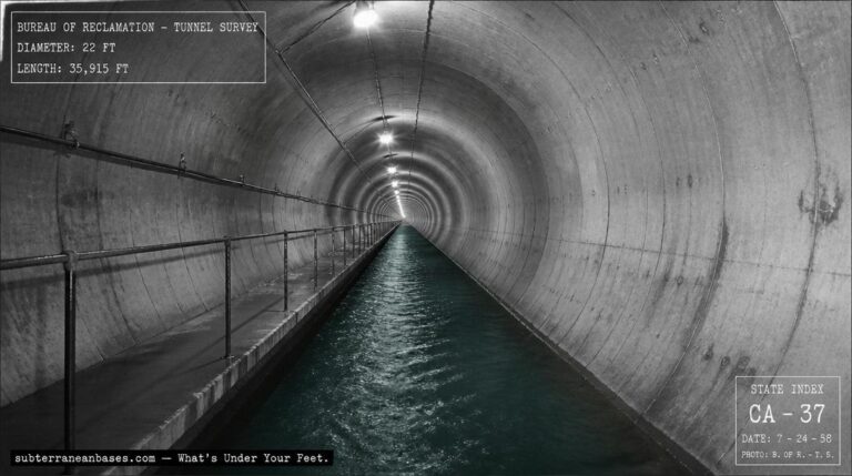The interior of a massive circular concrete-lined tunnel bored through mountain rock with a maintenance walkway and overhead lighting receding to a vanishing point and water flowing in a channel