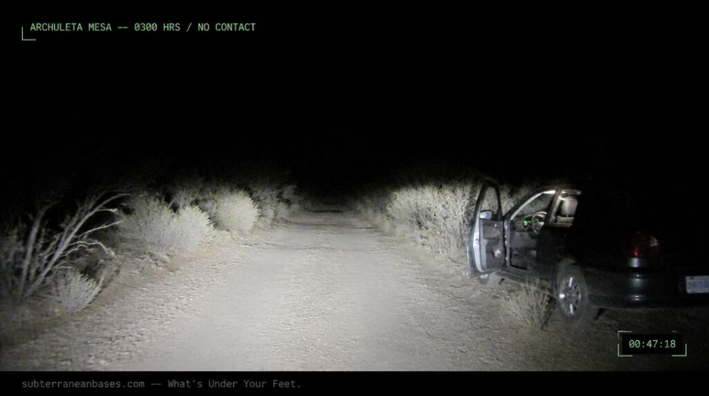 A narrow dirt road at night lit only by car headlights with scrubby desert brush and total darkness ahead and a parked car with door open at the roadside